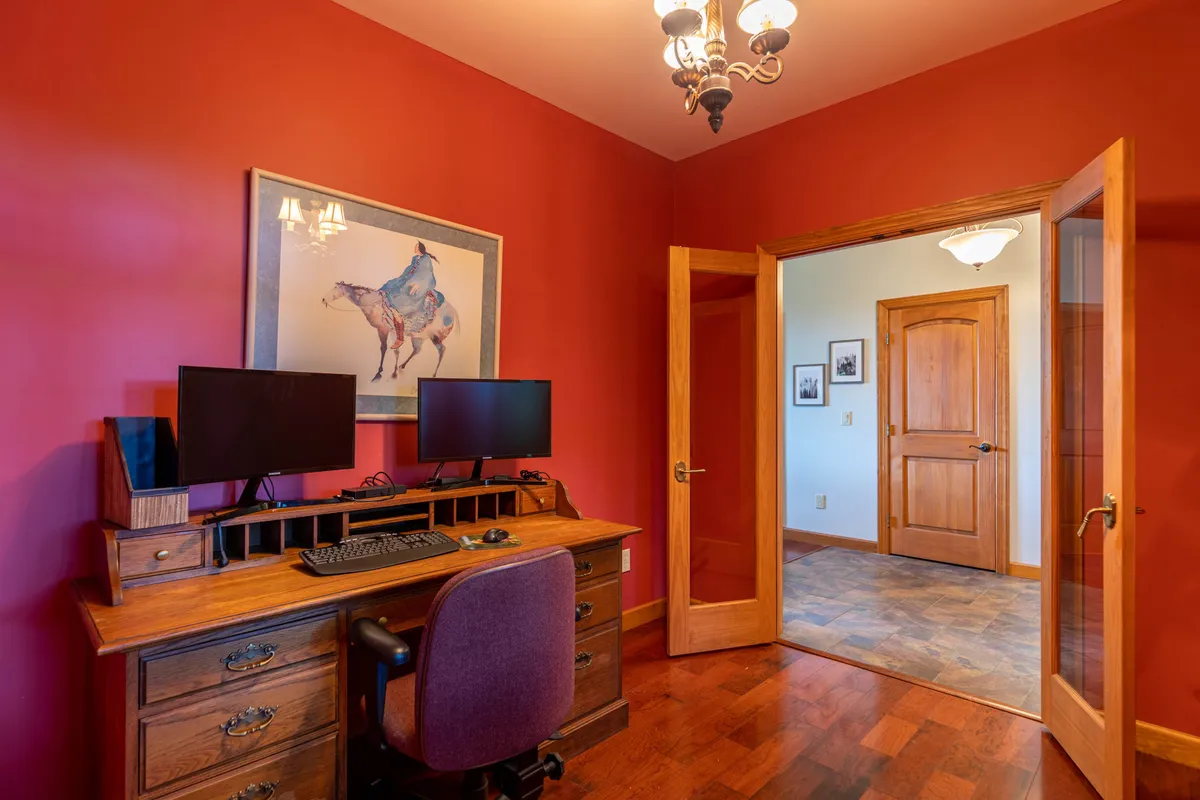 A dedicated office space featuring a wooden desk with dual monitors, tasteful artwork on the wall, and a warm chandelier overhead. The room exudes productivity and comfort, ideal for focused work sessions while enjoying the cozy mountain ambiance.