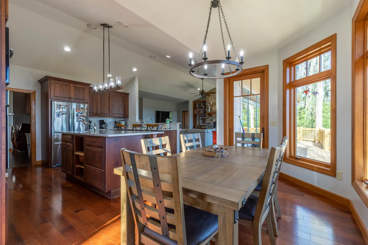 This image highlights the dining area with a central table, large windows that bring in natural light, a kitchen island in view, and a chandelier overhead. The atmosphere is inviting and set up for family meals, social gatherings, and celebrations.
