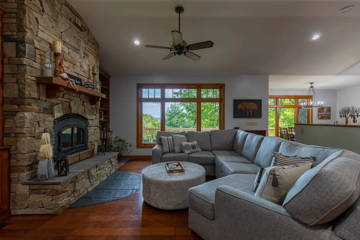 An inviting living area featuring a large sectional sofa, stone fireplace, and expansive windows with scenic views of the outdoors. The space is cozy and perfect for relaxing with family and pets after a day outside, while the windows provide a strong connection to the surrounding landscape.