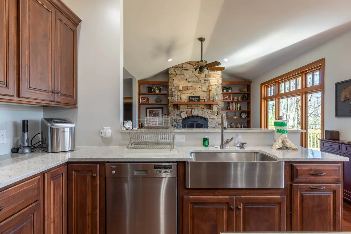 The image showcases a warm, inviting living area with a stone fireplace, wooden cabinetry, and expansive windows that let in natural light and reveal glimpses of the outdoors. The kitchen island and open space suggest a communal atmosphere perfect for gathering after days spent exploring the Blue Ridge Mountains.