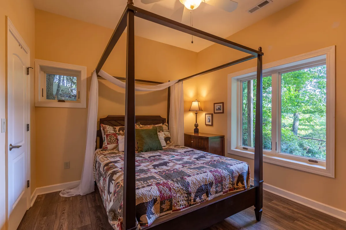 A serene bedroom with a four-poster bed, large window revealing glimpses of nature, a nightstand with a lamp, and a decorative quilt. The ambiance is both calming and inviting, with natural light streaming in and views of the mountains beyond.