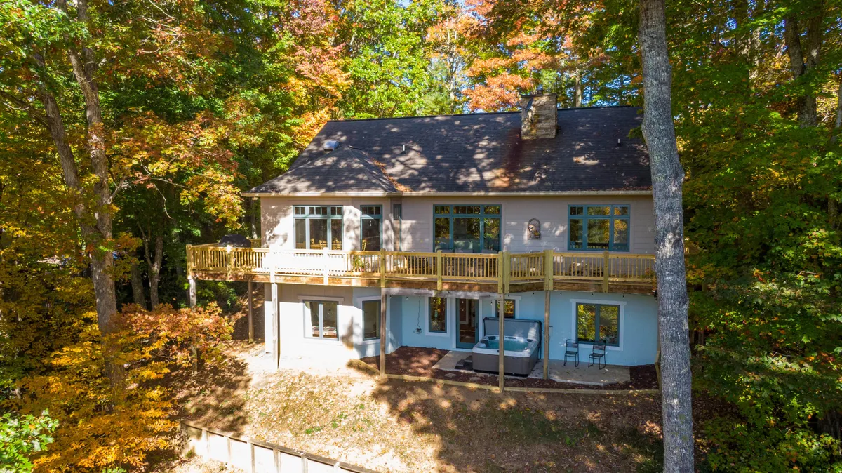 This image features a spacious wooden deck with comfortable seating and a patio area, framed by large windows and surrounded by lush trees. The natural setting offers tranquility and a sense of seclusion, while the deck is clearly furnished for outdoor dining and relaxation.