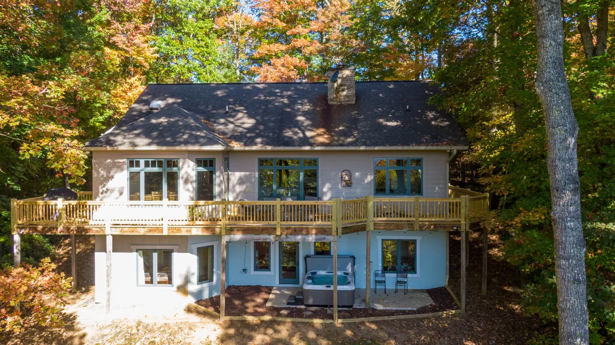 A view of the expansive wooden deck adorned with comfortable seating, a hot tub, and surrounded by lush trees. Large windows connect the indoor gathering spaces to the outdoors, and the serene natural backdrop is visible from the deck.
