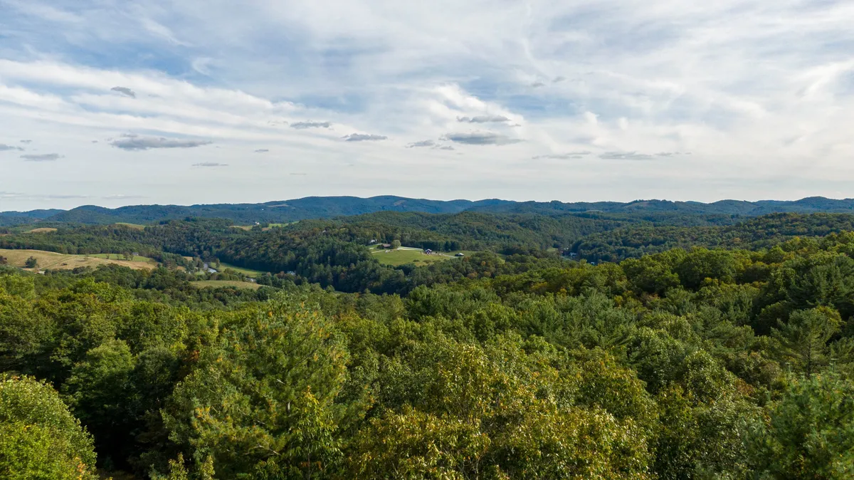 A sweeping view of rolling hills, dense tree cover, and distant mountains, providing a breathtaking natural panorama. The open fields and expansive sky evoke a sense of peace and connection with the surrounding landscape.