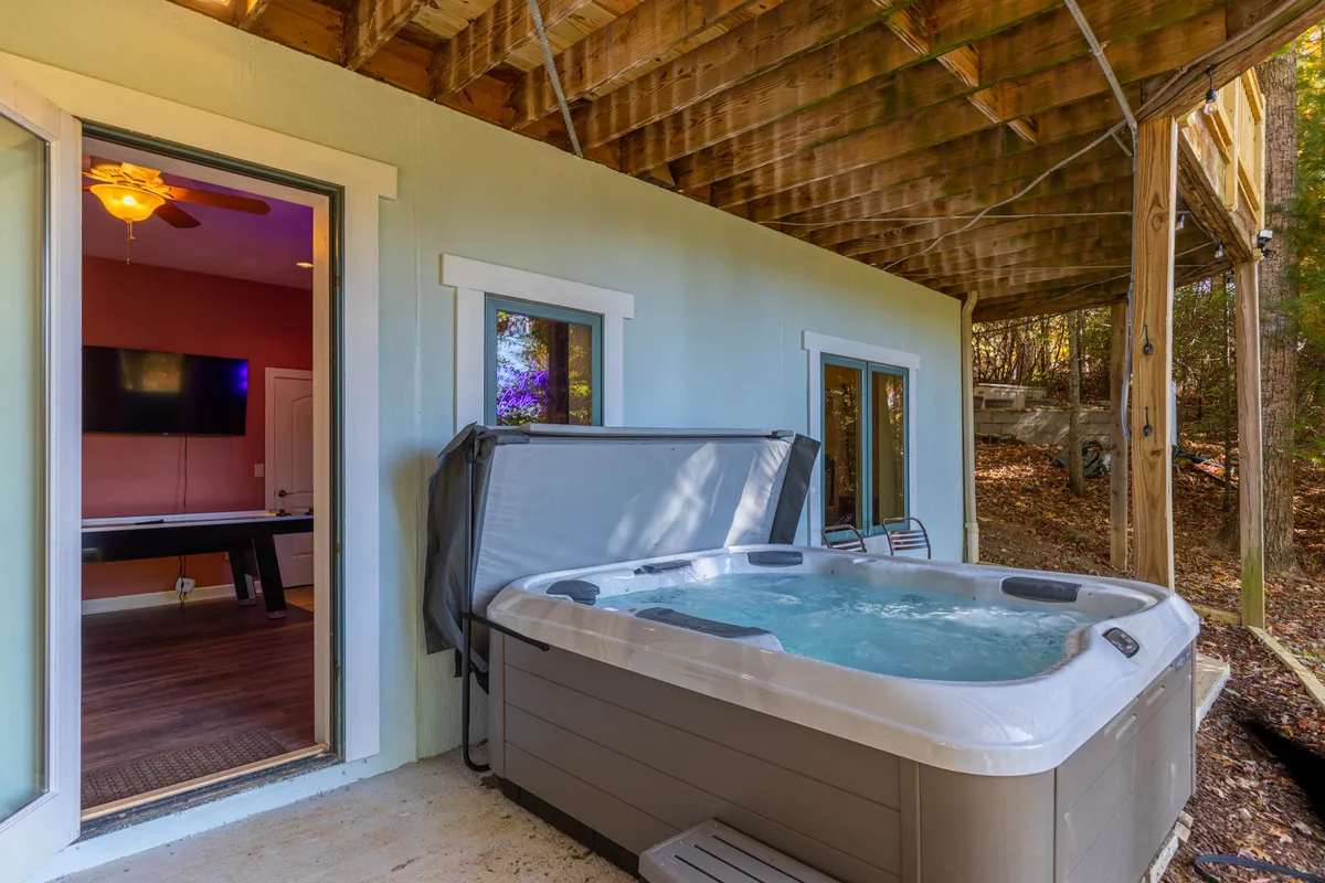 A tranquil exterior shot showing a hot tub nestled under a wooden structure, with open doors leading to the interior. The surrounding nature and relaxed setup invite multi-generational family enjoyment.