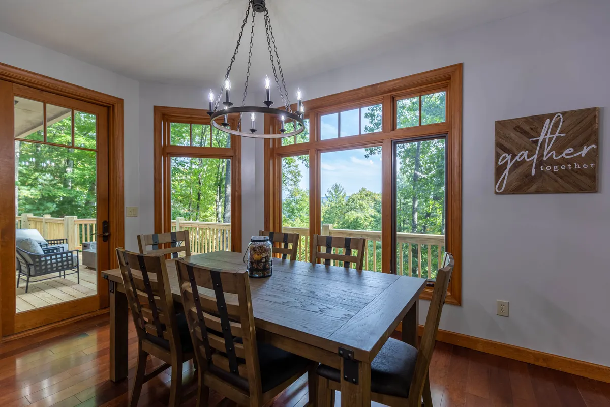 A cozy dining area featuring a large table ready for shared meals or game nights, flanked by large windows that offer sunset views and fill the space with natural light. Decor elements like the 'gather together' sign add extra warmth and invitation.