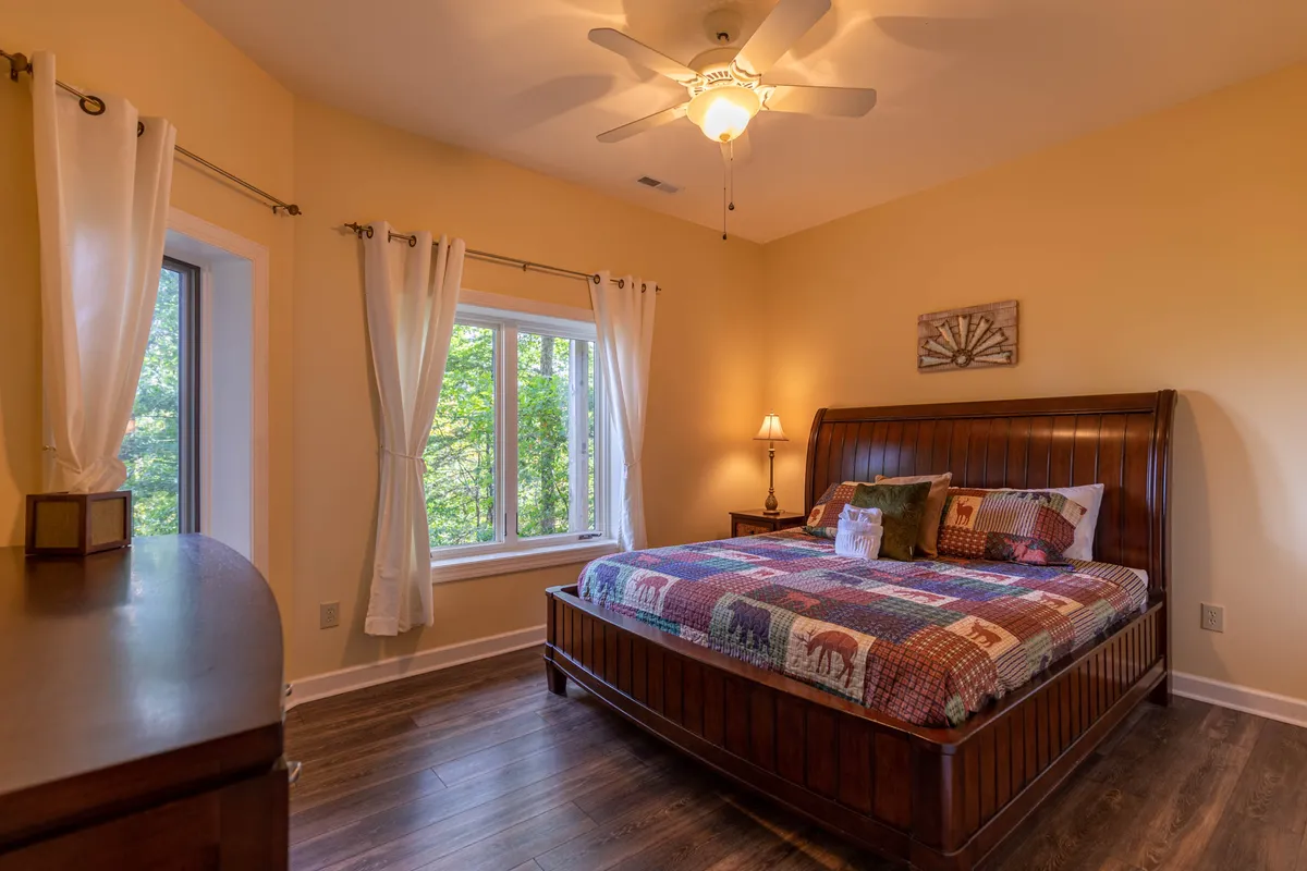 A welcoming bedroom featuring a bed positioned near a bright window, a nightstand, and a lamp. The view through the window suggests ever-changing seasonal scenery, inviting guests to appreciate the landscape from the comfort of the indoors.