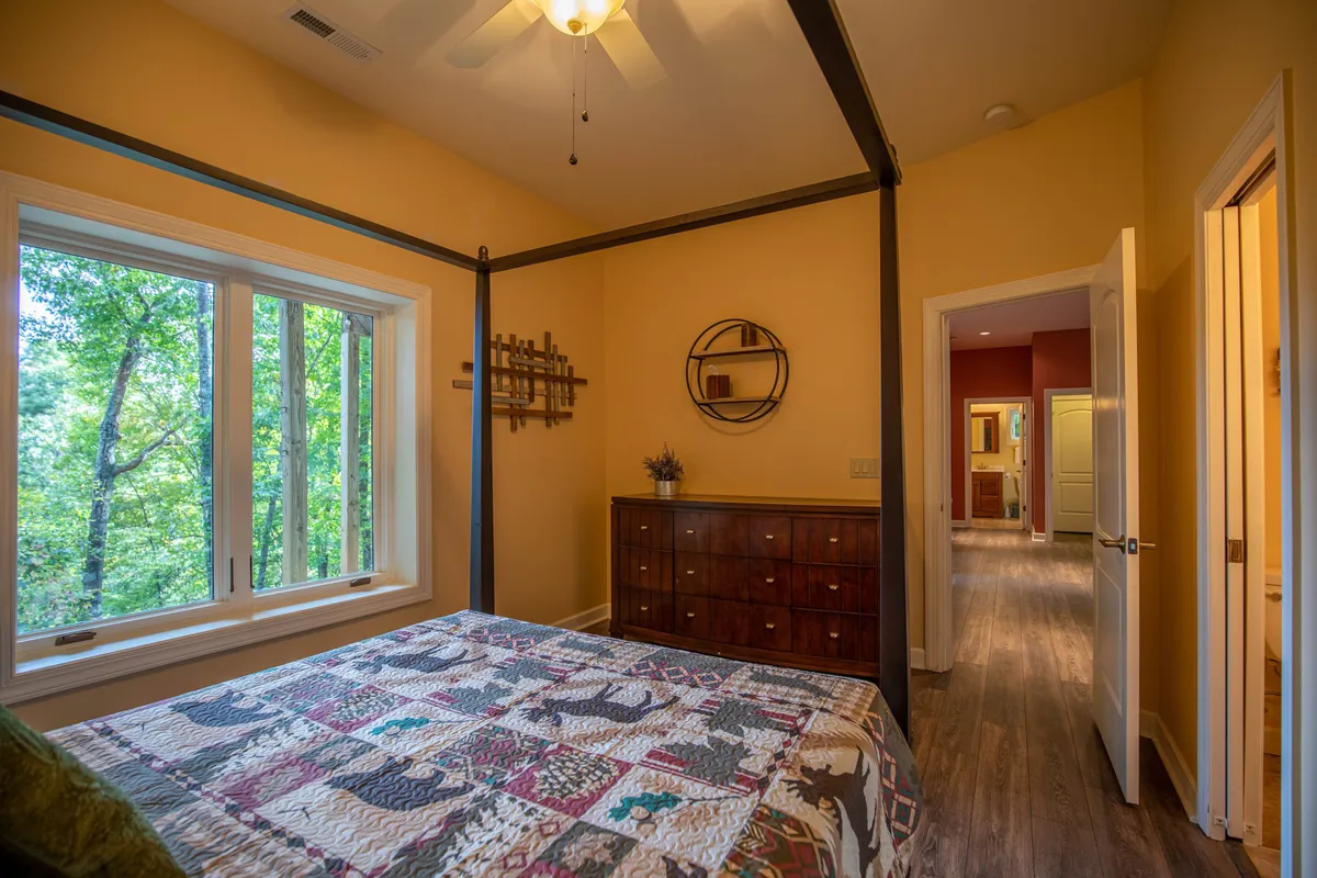 A serene master bedroom featuring a four-poster bed dressed in soft linens, large windows looking out to tranquil forest views, a wooden dresser, and cozy decorative touches. The space feels peaceful and is bathed in natural light, emphasizing a calm retreat atmosphere.